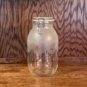 May include: A clear glass jar with a frosted upper half and a decorative snowflake design. The jar has a metal hinged lid with a clasp. The jar is sitting on a wooden surface against a wooden background.