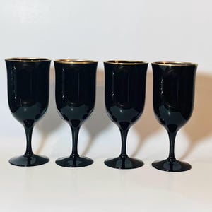 May include: Four black wine glasses with gold rims. The glasses have a classic goblet shape with a slender stem and a wide base. The glasses are arranged in a row against a white background.