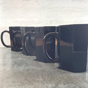 May include: Four black ceramic mugs with handles. The mugs are lined up in a row on a gray surface.