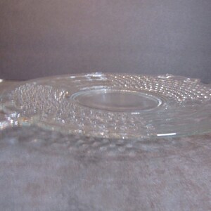 May include: A clear glass serving plate with a raised, textured design. The plate has a slightly scalloped edge and a center well.