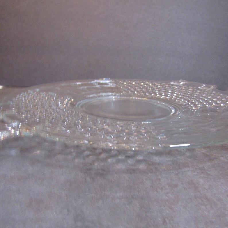 May include: A clear glass serving plate with a raised, textured design. The plate has a slightly scalloped edge and a center well.