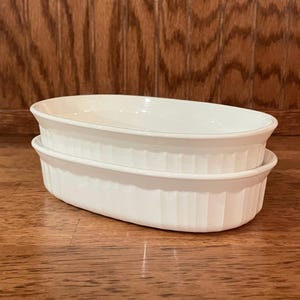 May include: Two white, oval-shaped ceramic baking dishes stacked on top of each other. The dishes have a ribbed design on the sides and a smooth, glossy finish. The dishes are on a wooden surface.