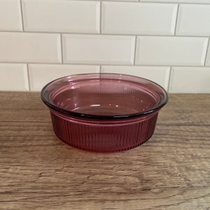 May include: A round, ribbed, clear glass bowl in a deep pink hue. The bowl has a slightly flared rim and sits on a wooden surface. The background features white rectangular tiles.
