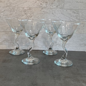 May include: Four clear martini glasses with a unique, swirled stem design. The glasses have a classic triangular bowl shape and sit on a round base. The background is a light gray brick wall.