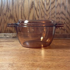 May include: A transparent amber-colored glass casserole dish with a matching lid. The round vessel features two integrated side handles and a smooth, polished finish, showcasing a classic design for kitchen use with a deep, wide basin.