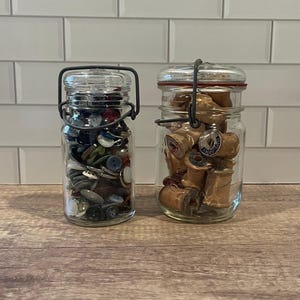 May include: Two glass jars with metal hinged lids. One jar is filled with a variety of buttons in different colors and sizes. The other jar contains wooden spools of thread, some with labels.