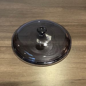 May include: A clear, round, glass lid with a dark gray tint. The lid features a central knob and a decorative, ridged pattern radiating from the knob. The lid is designed for cookware.