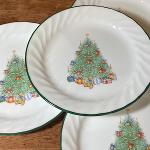 May include: Four white ceramic plates with a green trim and a spiral design. Each plate features a Christmas tree illustration with ornaments and presents. The plates are arranged on a wooden surface.