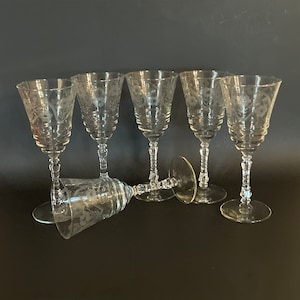 May include: Six clear, etched glass wine goblets with floral designs. The goblets have a classic shape with a flared bowl, a slender stem, and a round base. One goblet is lying on its side. The background is a solid black.