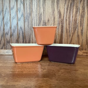 May include: Three ceramic baking dishes, one orange, one light orange, and one purple. The dishes are stacked on top of each other.