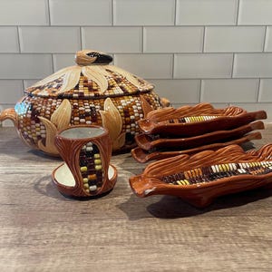 May include: A ceramic corn-themed serving set. The set includes a lidded serving dish, a small cup, and several corn-shaped serving dishes. The pieces are primarily brown, yellow, and white, with detailed textures.