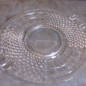 May include: A clear glass serving dish with a raised, textured design. The dish has two handles on the sides and a raised center section.