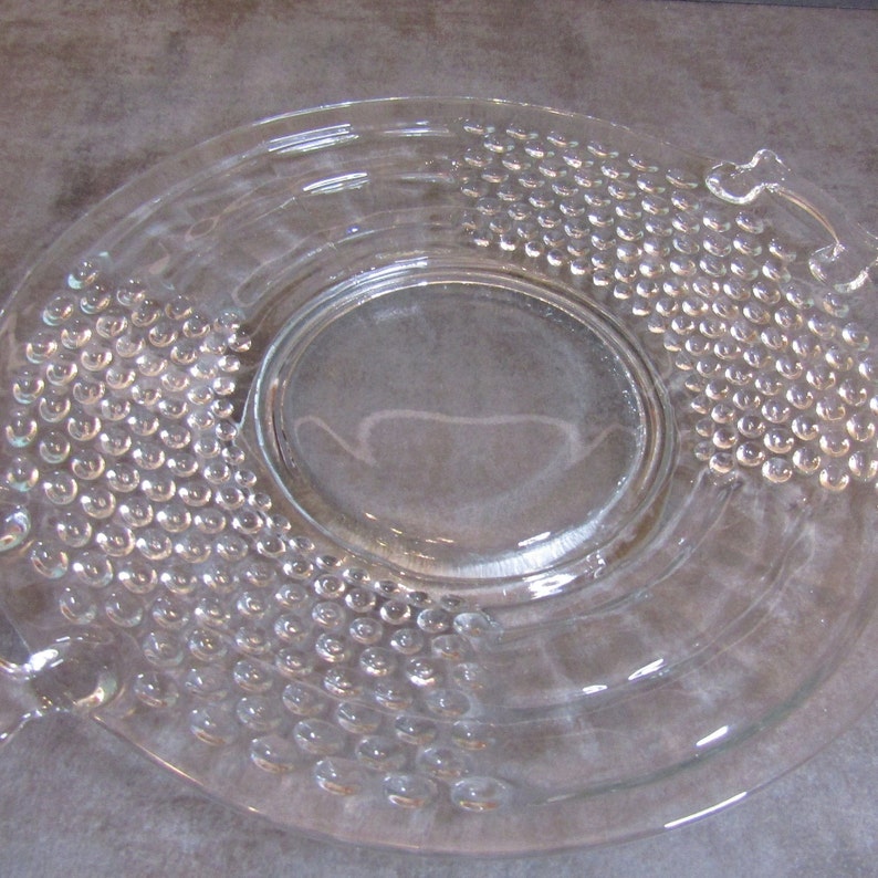 May include: A clear glass serving dish with a raised, textured design. The dish has two handles on the sides and a raised center section.