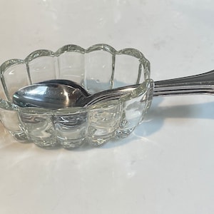 May include: A clear glass dish with a scalloped edge, holding several silver spoons. The spoons are neatly arranged, with their handles extending out of the dish. The dish and spoons are on a white surface.