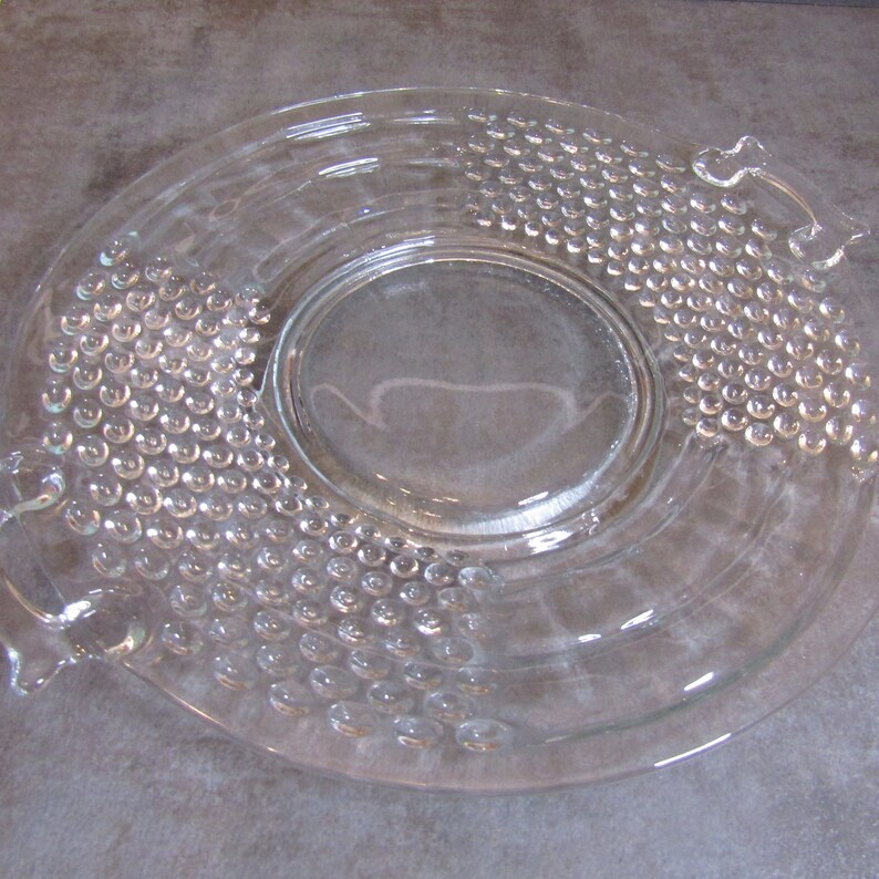 May include: A clear glass serving dish with a raised, textured pattern of small circles. The dish has two handles on the sides.