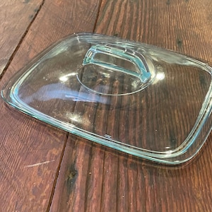 May include: A clear, rectangular glass lid with a rounded handle. The lid has a slightly teal-colored trim and handle, and is designed for cookware. The lid is resting on a wooden surface.