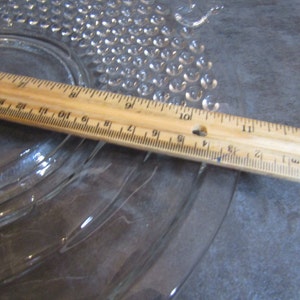 May include: A clear glass plate with a textured surface. The plate has a raised, scalloped edge and a series of concentric circles. A wooden ruler is placed on top of the plate, with the numbers 7 through 12 visible.