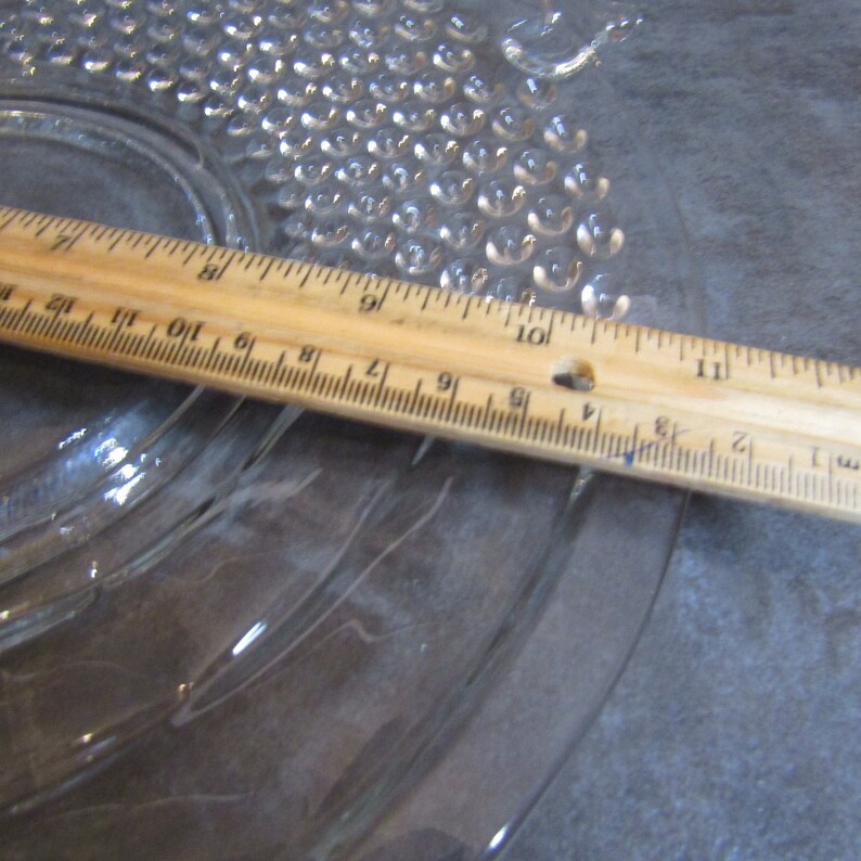 May include: A clear glass plate with a textured surface. The plate has a raised, scalloped edge and a series of concentric circles. A wooden ruler is placed on top of the plate, with the numbers 7 through 12 visible.