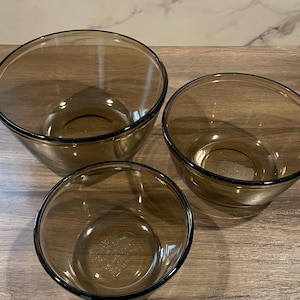 May include: Set of three amber-colored glass mixing bowls. The bowls are nested, with a graduated size difference. Each bowl has a dark rim. The bowls are made of clear glass and are sitting on a wooden surface.