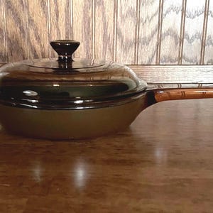 May include: A vintage olive green saucepan with a glass lid and a dark brown handle. The lid has a dark brown knob. The pan is sitting on a wooden surface. The background is a wooden wall.