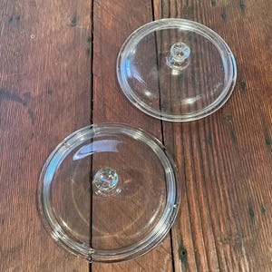 May include: Two clear glass pot lids with small, round knobs. The lids are circular with a slightly raised rim. The knobs are also clear and round. The lids are on a dark brown wooden surface.