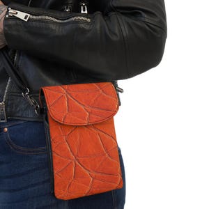 May include: A small orange leather crossbody bag with a flap closure and a textured pattern. The bag is worn by a person wearing a black leather jacket and blue jeans.