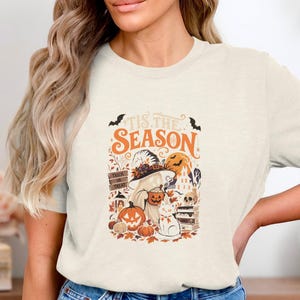 May include: A cream-colored short-sleeve t-shirt featuring a graphic print with the text 'Tis the Season'. The design includes a ghost wearing a witch hat, a cat, pumpkins, a skull, books, and bats in orange, black, and brown tones.