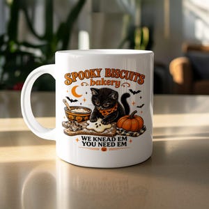 May include: A white ceramic mug featuring a graphic of a black kitten wearing an orange bandana, baking ghost-shaped cookies. The text reads SPOOKY BISCUITS bakery and WE KNEAD EM YOU NEED EM, accompanied by illustrations of a pumpkin, bats, and a crescent moon.