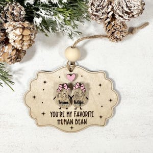 Puede incluir: Un adorno de madera con borde festoneado, con dos personajes de dibujos animados de frijoles con lazos y los nombres "Ivana" y "Lidija". Un corazón rosa se encuentra encima de los frijoles, y el texto "YOU'RE MY FAVORITE HUMAN BEAN" está debajo. El adorno cuelga de una cuerda.