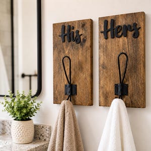 May include: Two rectangular wooden wall plaques with a dark brown finish, each featuring a black metal hook. The left plaque has the word His in black script, and the right plaque has the word Hers in black script. A beige towel hangs on the left hook, and a white towel hangs on the right hook.