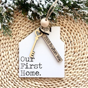 May include: White wooden house-shaped ornament with a key and heart charm. The ornament says "Our First Home" and "2008 Bradham Way" and "2024".