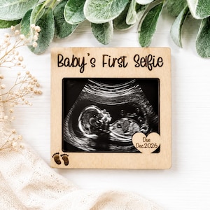 May include: A square light-coloured wooden frame holding a black and white ultrasound image. The top of the frame is engraved with the text "Baby's First Selfie." A small heart-shaped cutout displays "Due Dec.2026," and two small footprints are engraved.