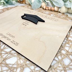 May include: A wooden graduation photo frame with the words "Little Graduate" and "your photo here" engraved. A black mortarboard is affixed to the frame. The name "Lincoln" and the year "2025" are also engraved.