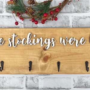 May include: Wooden sign with the phrase "and the stockings were hung..." in white script. The sign has six black metal hooks and is decorated with a small wreath with red berries.