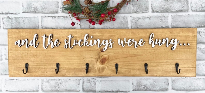 May include: Wooden sign with the phrase "and the stockings were hung..." in white script. The sign has six black metal hooks and is decorated with a small wreath with red berries.