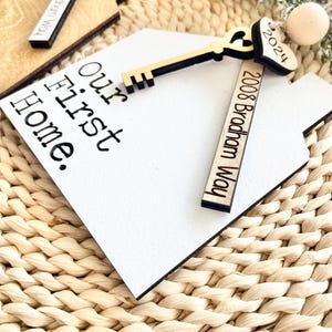 May include: White wooden house-shaped ornament with the text "Our First Home" and a gold key with a heart-shaped keyhole. The ornament also includes a wooden tag with the text "2008 Bradham Way" and a small heart-shaped tag with the text "2024".