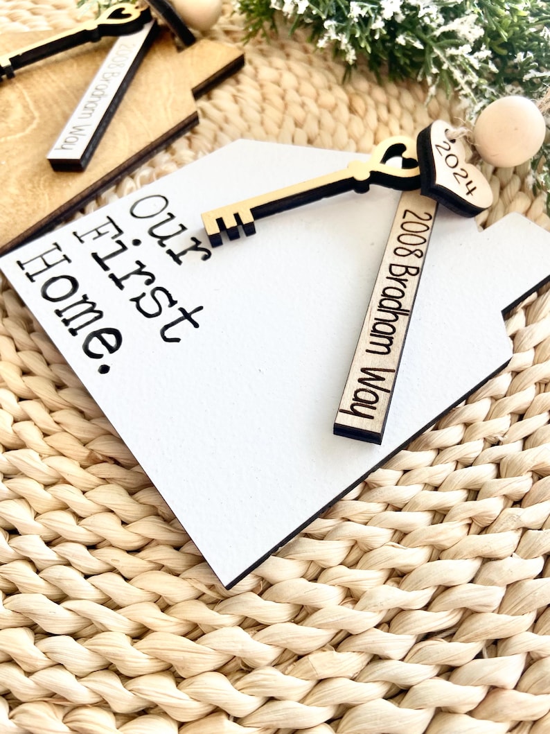 May include: White wooden house-shaped ornament with the text "Our First Home" and a gold key with a heart-shaped keyhole. The ornament also includes a wooden tag with the text "2008 Bradham Way" and a small heart-shaped tag with the text "2024".