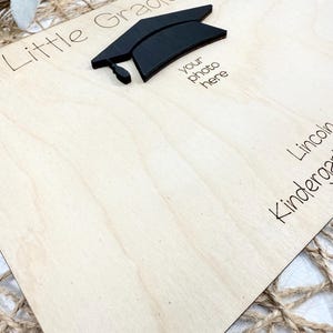 May include: A wooden graduation photo frame with the words "Little Graduate" and "your photo here" printed on it. A black graduation cap is affixed to the frame. The frame is light-colored wood.