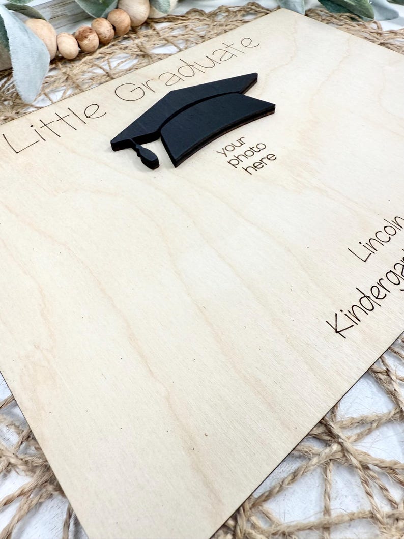 May include: A wooden graduation photo frame with the words "Little Graduate" and "your photo here" printed on it. A black graduation cap is affixed to the frame. The frame is light-colored wood.