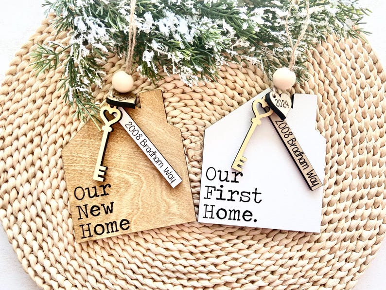May include: Two wooden house-shaped Christmas ornaments.  One is light brown with the text 'Our New Home' and address '2008 Bradham Way'; the other is white with 'Our First Home.' and the same address. Both feature a small key charm.  The ornaments are displayed against a woven background.