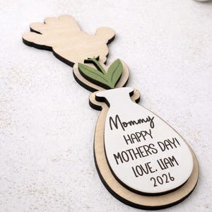 Op de afbeelding: Houten Moederdagdecoratie in de vorm van een vaas, hand en bladeren. De vaas heeft de tekst "Mommy HAPPY MOTHERS DAY! LOVE. LIAM 2026" in het zwart. De decoratie is gemaakt van lichtgekleurd hout met zwarte accenten.