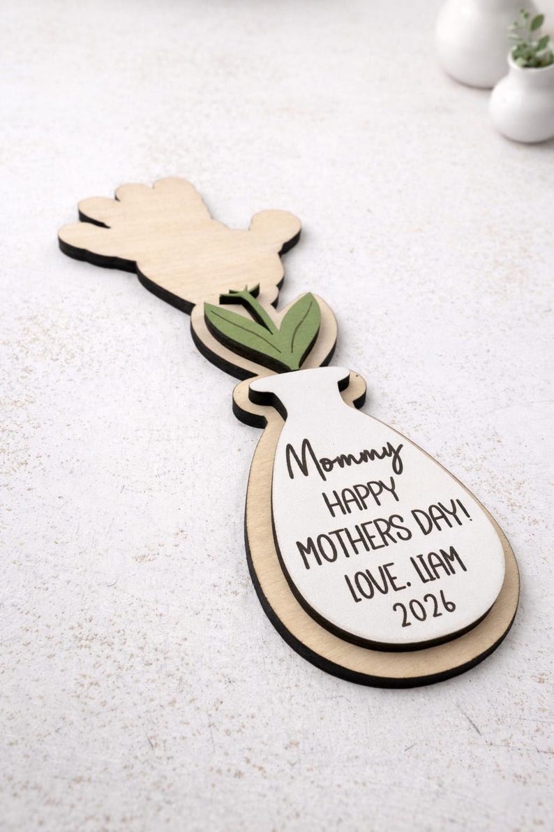Op de afbeelding: Houten Moederdagdecoratie in de vorm van een vaas, hand en bladeren. De vaas heeft de tekst "Mommy HAPPY MOTHERS DAY! LOVE. LIAM 2026" in het zwart. De decoratie is gemaakt van lichtgekleurd hout met zwarte accenten.