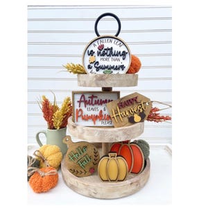 May include: A three-tiered wooden tray with a black metal handle on the top tier. The tray is decorated with fall-themed signs and pumpkins. The signs read "A fallen leaf is nothing more than a summer's...", "Autumn leaves & pumpkin please", and "Happy Harvest".
