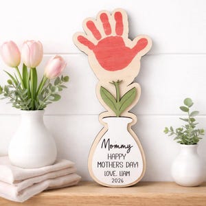 Op de afbeelding: Houten Moederdag bloemdecoratie met een rode handafdruk, groene bladeren en een witte vaasvorm. De tekst luidt "Mommy, Happy Mother's Day! Love, Liam 2026".