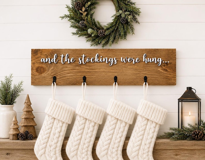 May include: A wooden sign with the text "and the stockings were hung..." above four white cable-knit Christmas stockings. A green wreath and a lit candle in a black lantern add to the holiday decor.