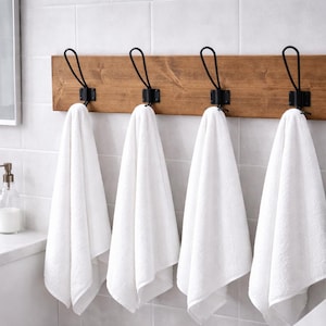 May include: A wooden towel rack with four black metal hooks, each holding a folded white towel. The rack is mounted on a tiled wall, with a sink and mirror visible in the background. A small plant in a white pot is on the right.