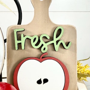 Op de afbeelding: Een lichtbruin houten bord in de vorm van een snijplank met het woord Fresh in lichtgroene schrijfletters en een afbeelding van een doorgesneden appel met een rode rand, wit midden en twee zwarte pitten onder de tekst.