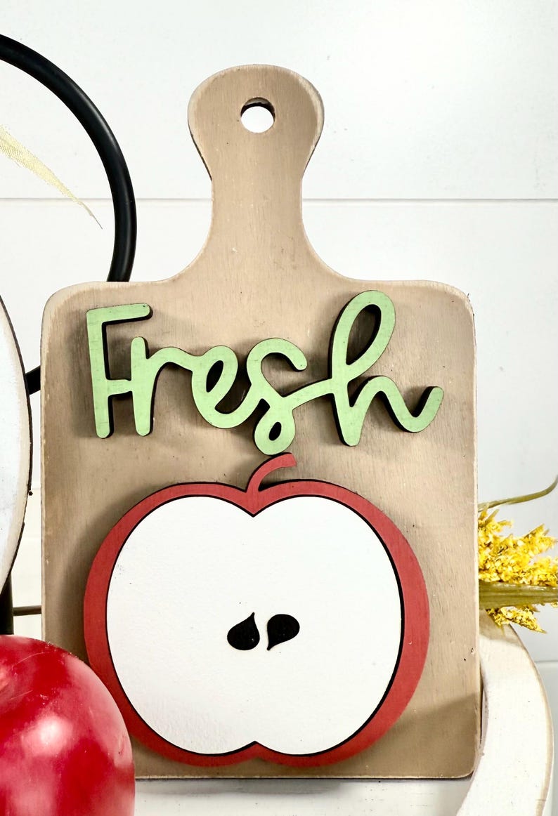 Op de afbeelding: Een lichtbruin houten bord in de vorm van een snijplank met het woord Fresh in lichtgroene schrijfletters en een afbeelding van een doorgesneden appel met een rode rand, wit midden en twee zwarte pitten onder de tekst.