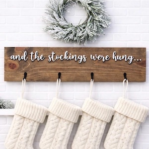 May include: A wooden sign with the phrase "and the stockings were hung..." in white script. Below the sign are four black hooks holding white cable-knit Christmas stockings. A snowy wreath and greenery decorate the background.