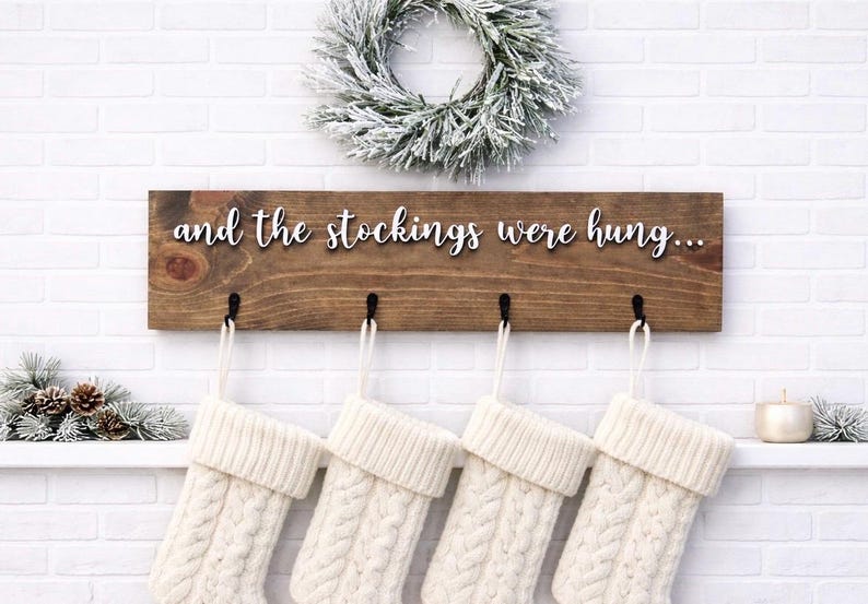 May include: A wooden sign with the phrase "and the stockings were hung..." in white script. Below the sign are four black hooks holding white cable-knit Christmas stockings. A snowy wreath and greenery decorate the background.